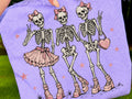 Light purple graphic tee with skeleton girl, skull faces, bows, and stars