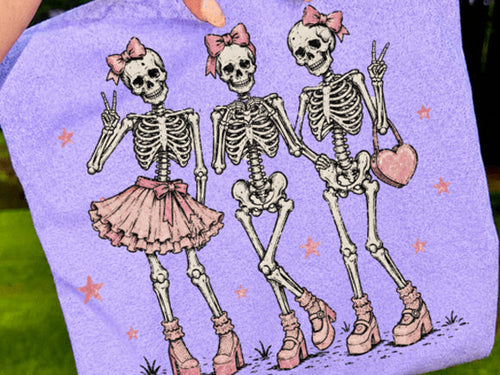 Light purple graphic tee with skeleton girl, skull faces, bows, and stars