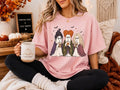 Pink graphic tee with three cartoon witches holding coffee mugs and star accents
