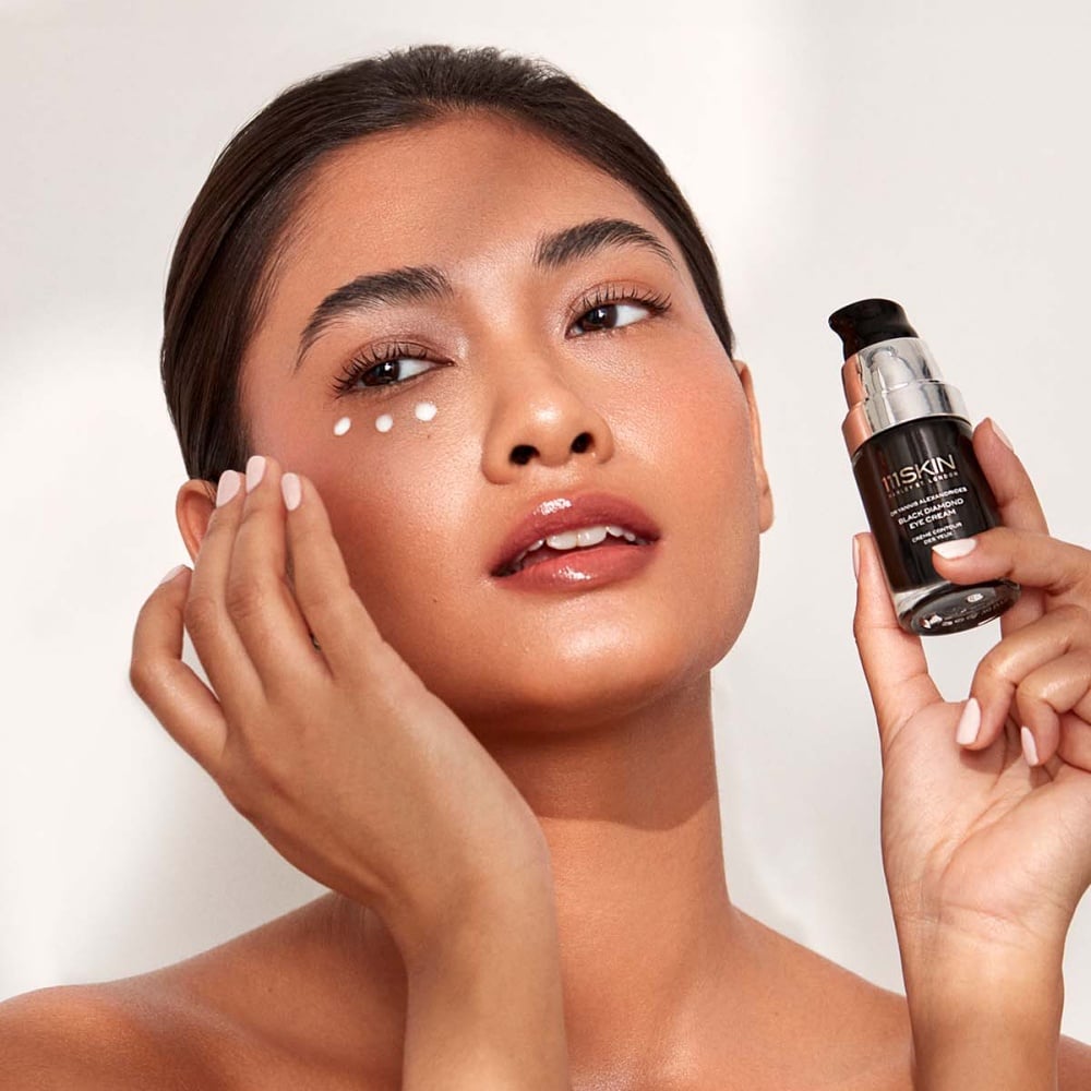 Woman applying eye cream near one eye with a small black skincare bottle