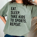 Moss green unisex graphic tee with black text: EAT SLEEP TAKE KIDS TO SPORTS REPEAT.