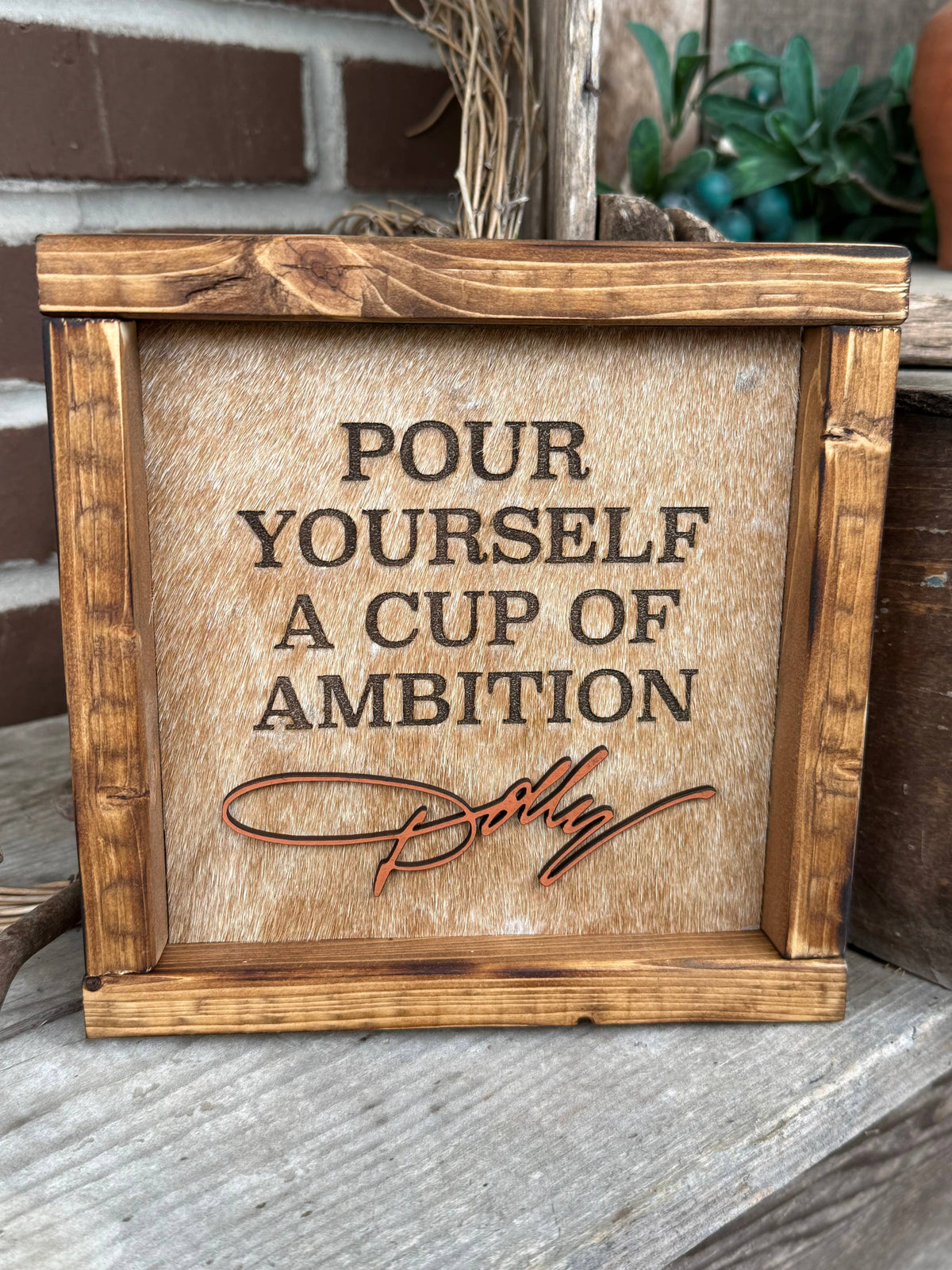 Framed cowhide sign on tabletop, 'POUR YOURSELF A CUP OF AMBITION' in serif font