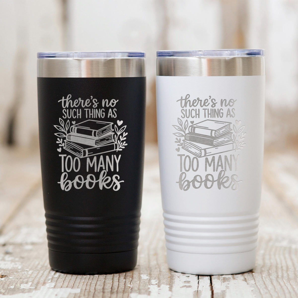 Two insulated travel mugs on wood: black and white, each with “Too many books” quote