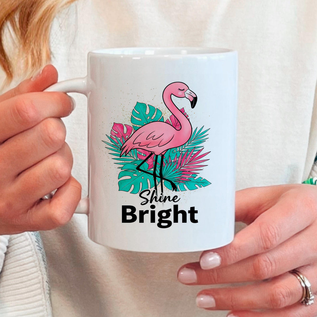 Hand holding white mug featuring pink flamingo, tropical leaves, and “Shine Bright”