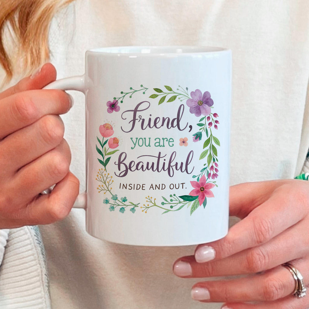 Hand holding white mug with floral wreath and “Friend, you are Beautiful” saying