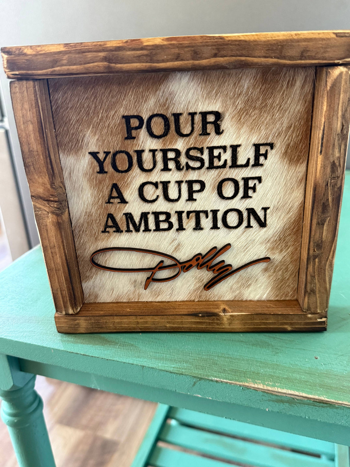 Framed cowhide wall sign with 'POUR YOURSELF A CUP OF AMBITION' text and signature