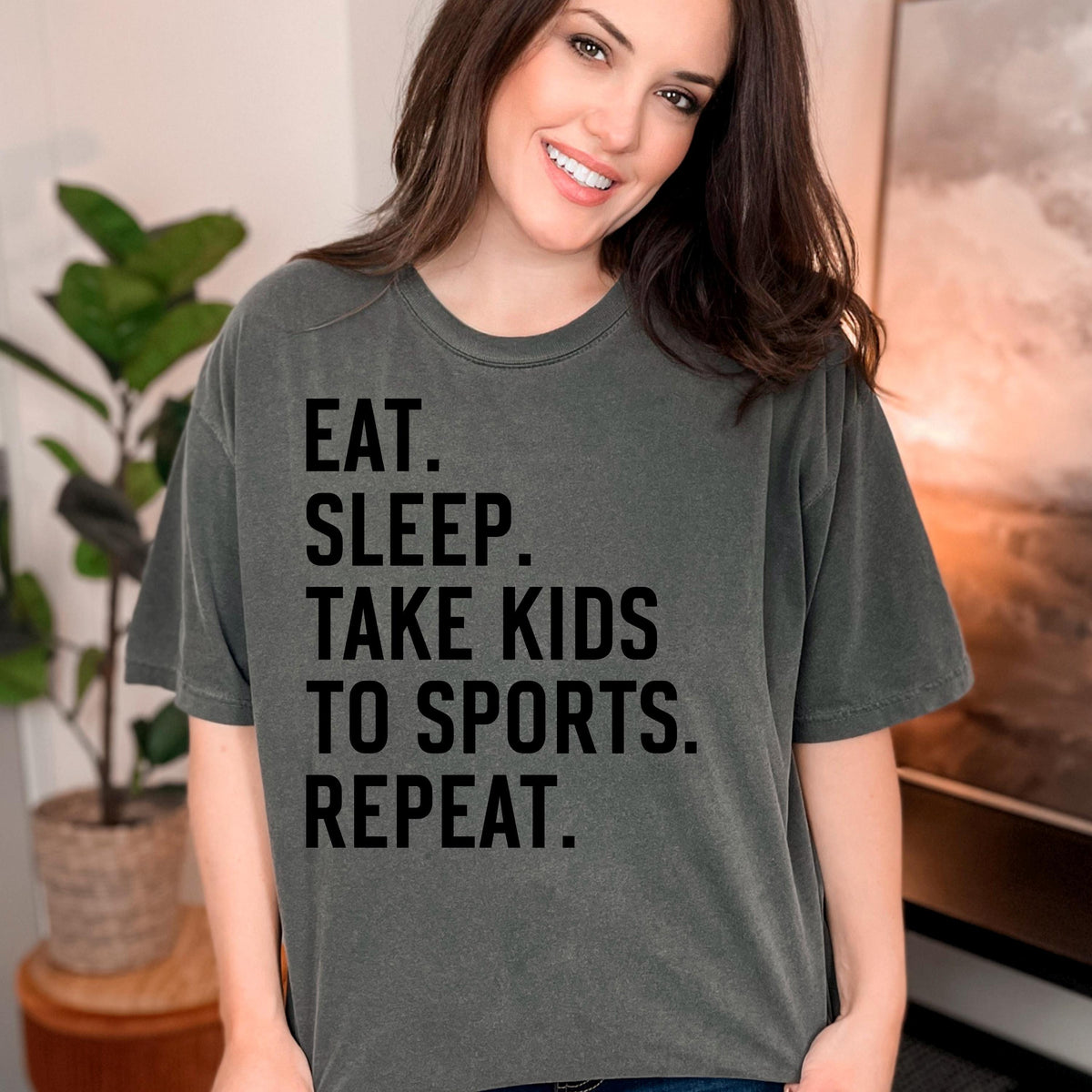 Olive unisex graphic tee with black text: EAT SLEEP TAKE KIDS TO SPORTS REPEAT.