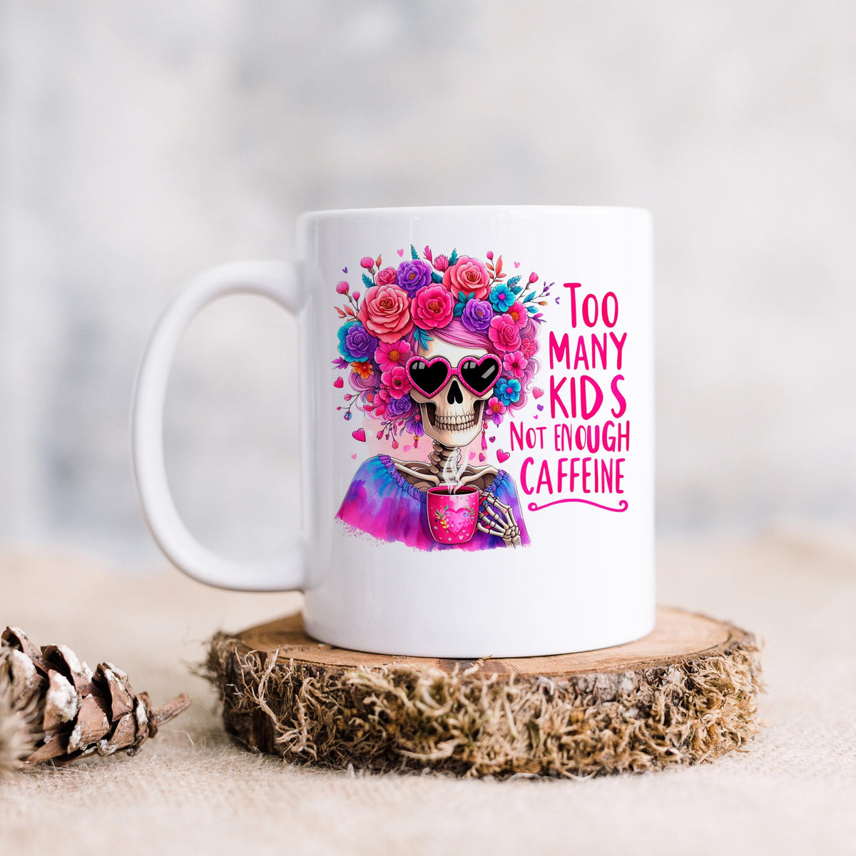 White ceramic mug on a wood base with floral crown skull and “Too Many Kids” coffee message.