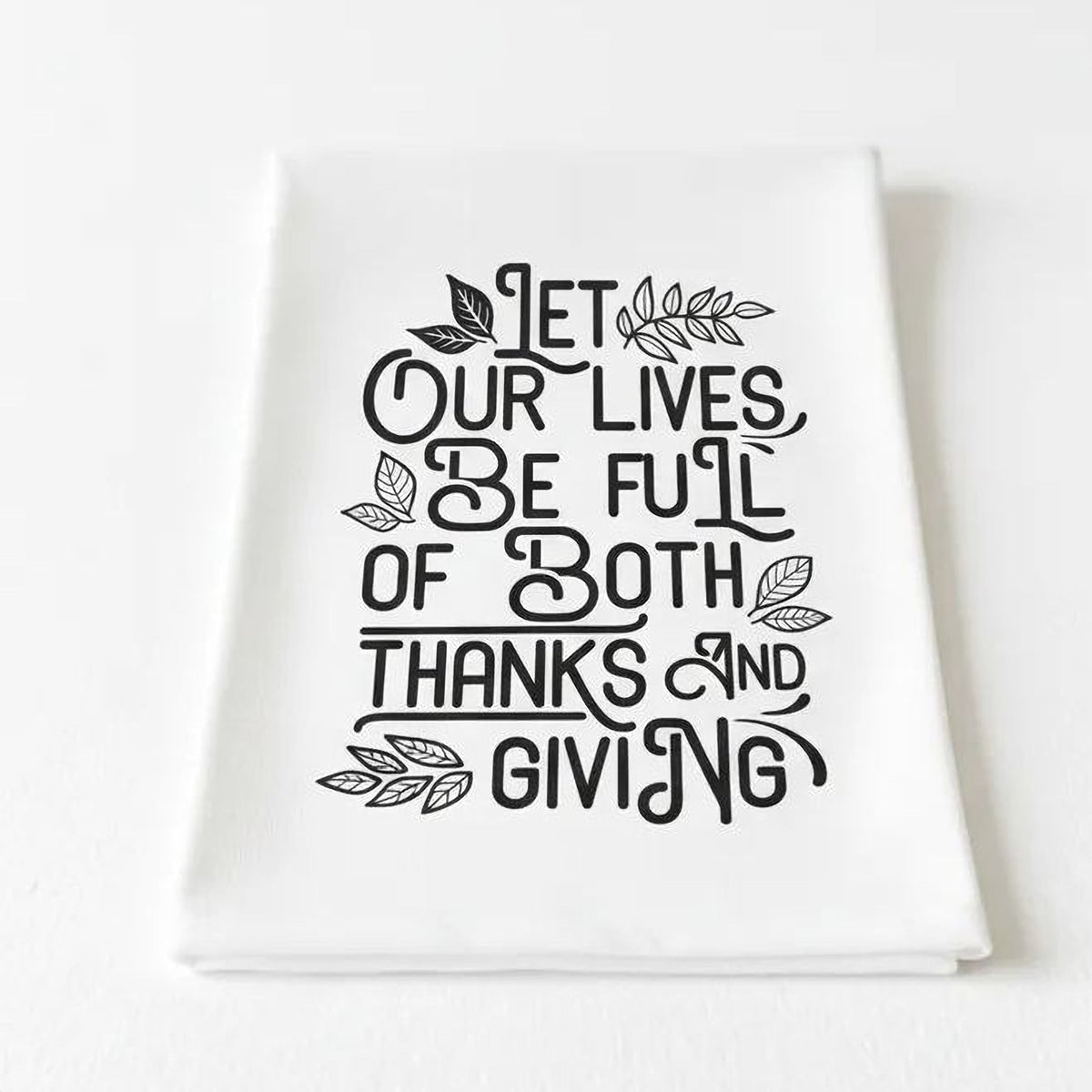 White Thanksgiving kitchen towel folded, featuring black “Let our lives be full of both…”
