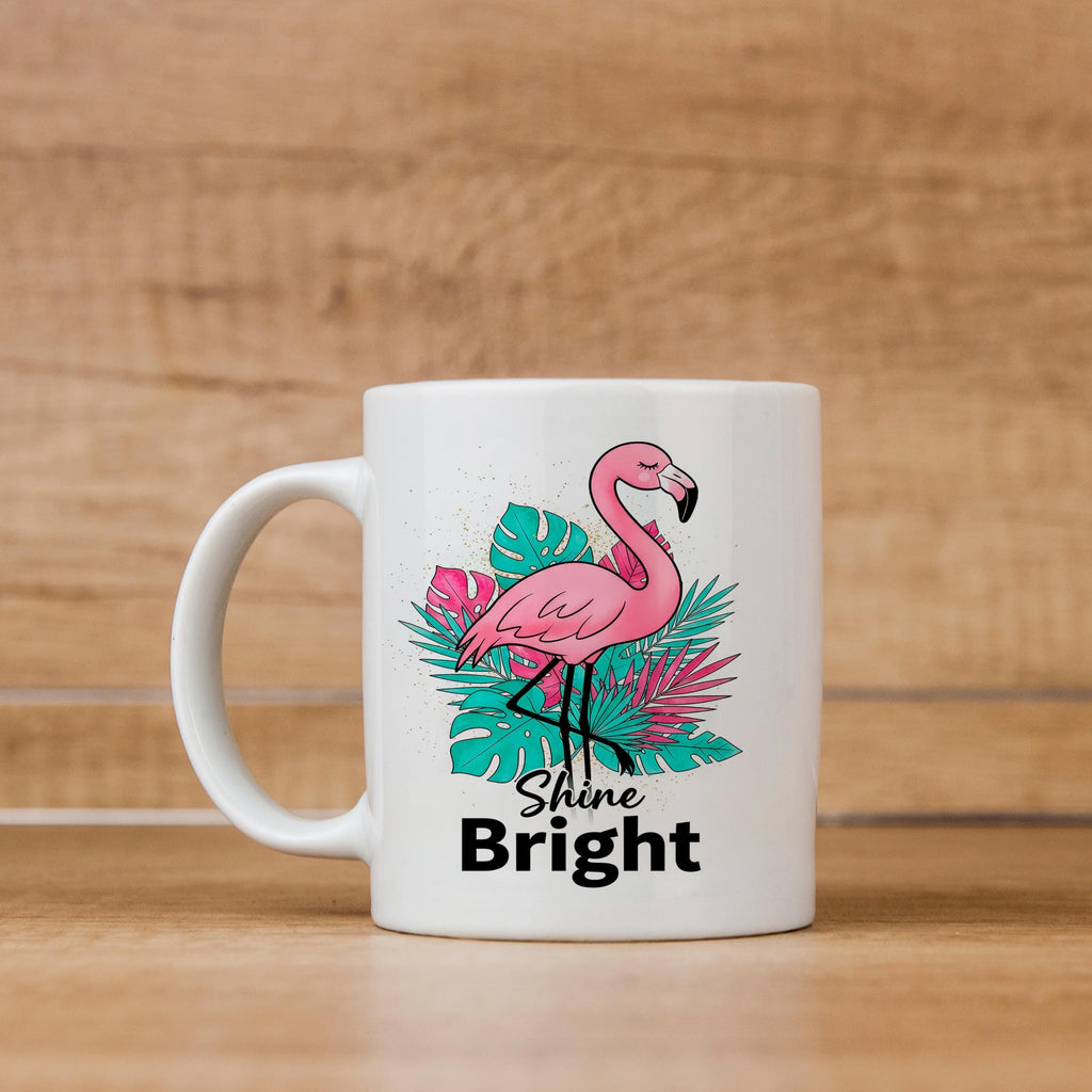 White ceramic mug with pink flamingo on tropical leaves and “Shine Bright” text