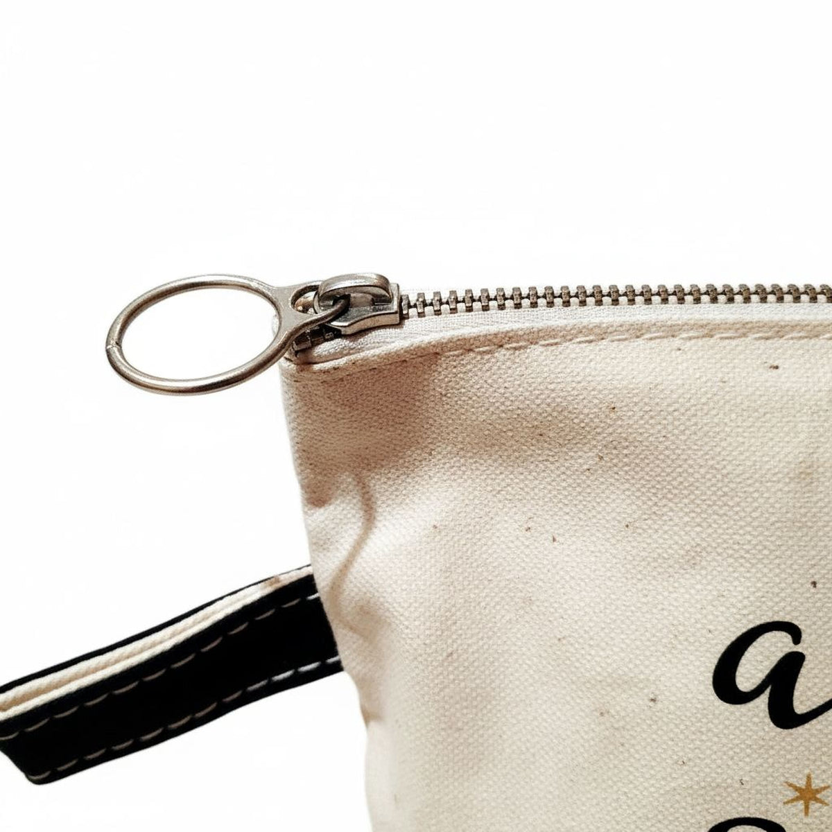Beige canvas pouch close-up showing metal zipper pull and black trim seam.