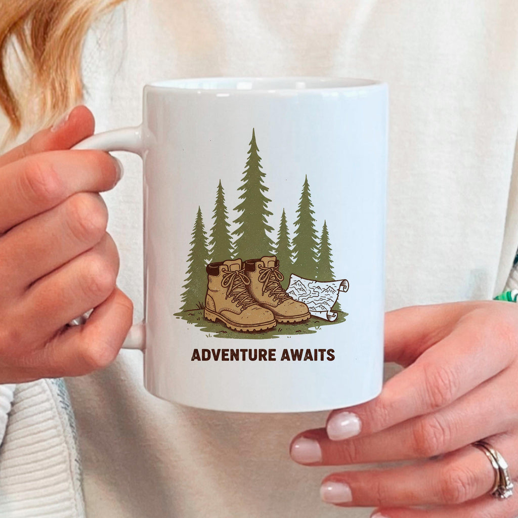 White ceramic mug held in hands, 'ADVENTURE AWAITS' boots and pine trees graphic