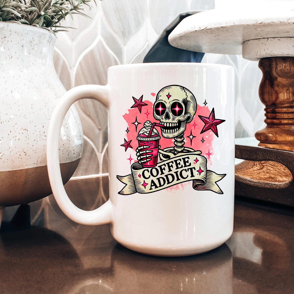 White ceramic coffee mug with pink skull 'Coffee Addict' graphic, on a table beside decor