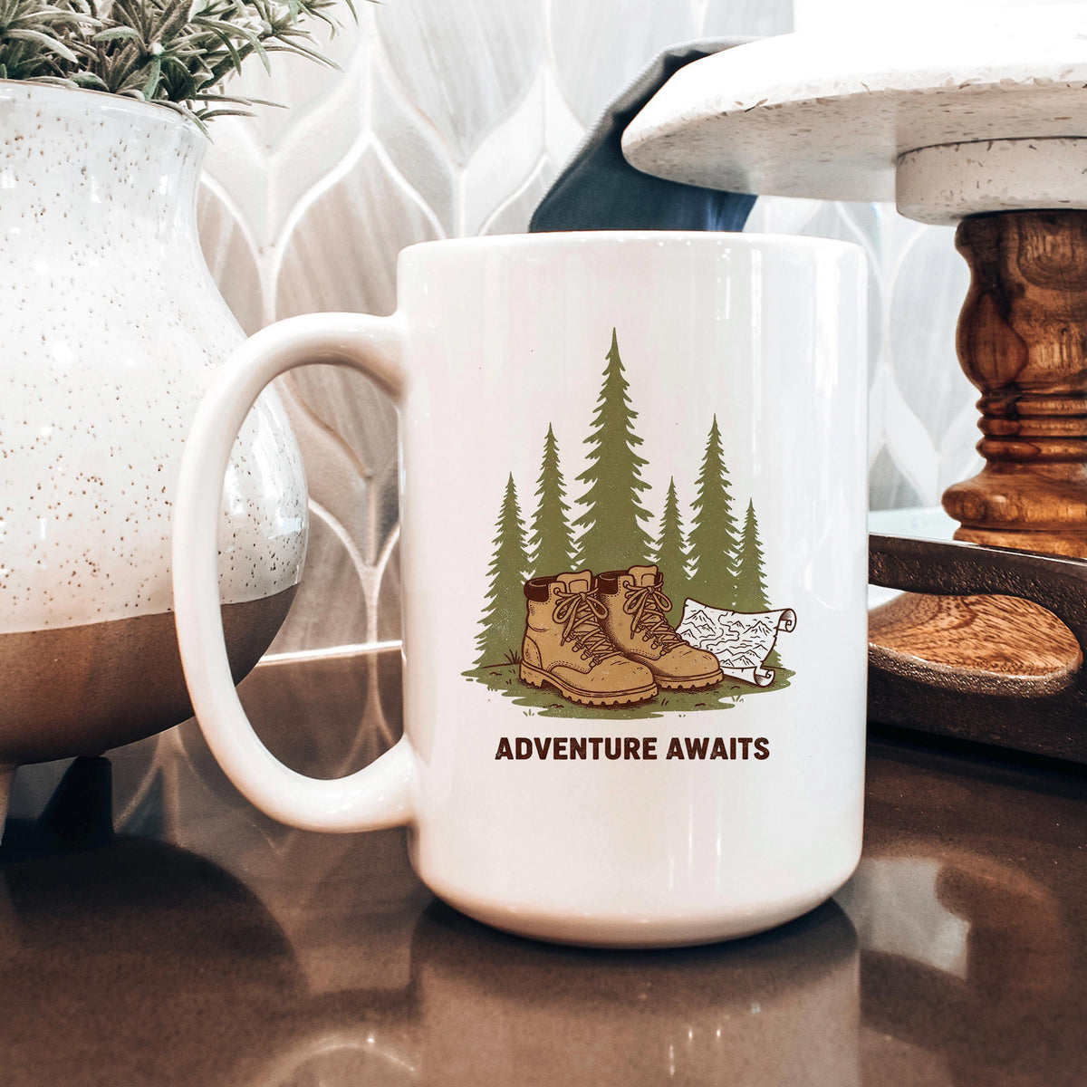 White ceramic mug, angled view, with 'ADVENTURE AWAITS' boots and pine trees graphic