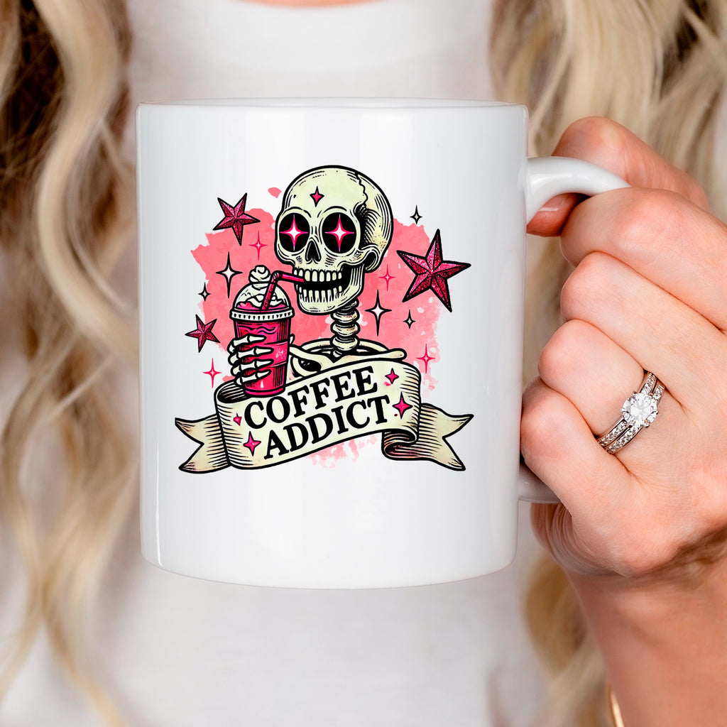 White ceramic coffee mug with pink skull and 'Coffee Addict' graphic, held in a hand