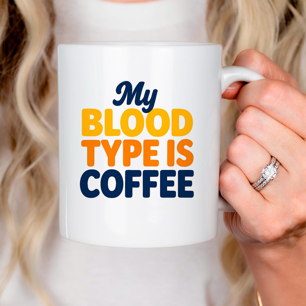 White ceramic coffee mug being held near face, “My BLOOD TYPE IS COFFEE” printed.