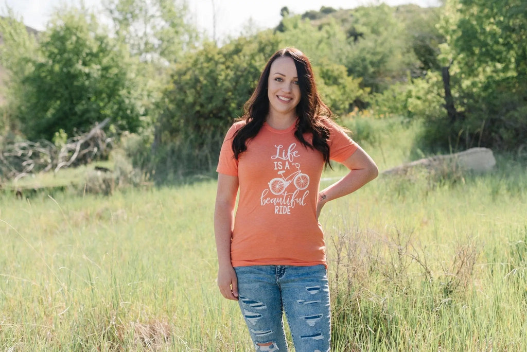 Coral graphic bicycle quote T-shirt on model, featuring white “Life is a beautiful ride”