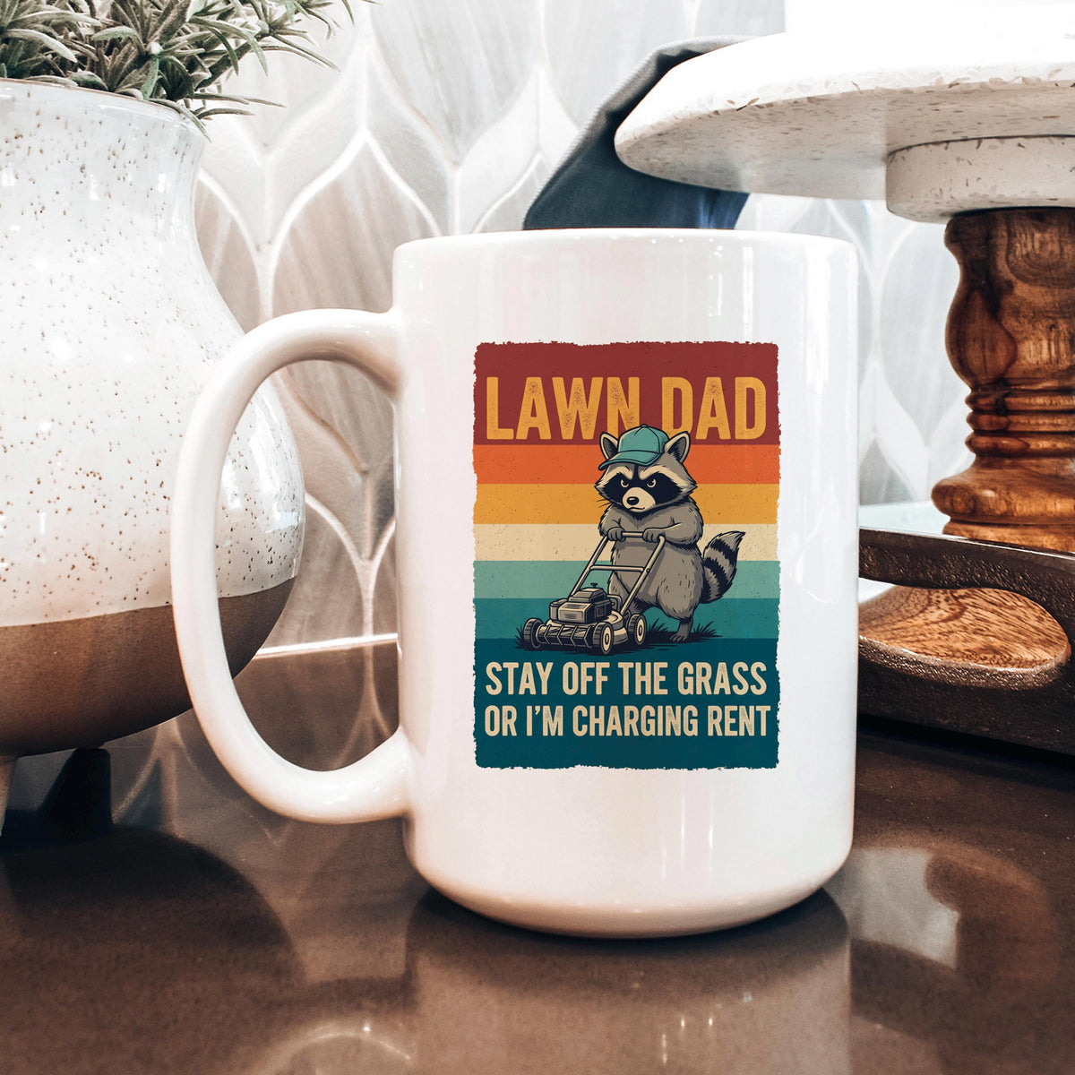 White coffee mug with raccoon mowing graphic and “OR I'M CHARGING RENT” text