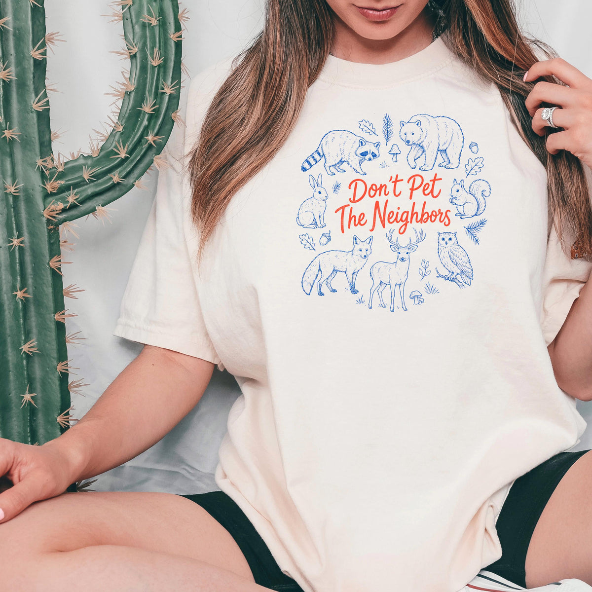 Ivory T-shirt close-up with blue animal woodcut and red 'Don't Pet The Neighbors' text