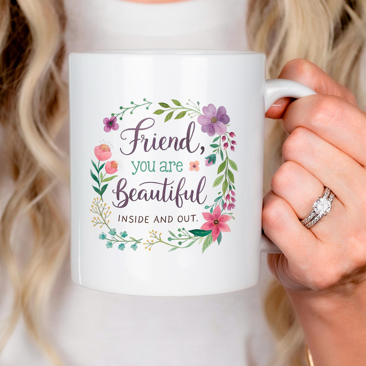 Person holding white mug with floral wreath and “Friend, you are Beautiful” text