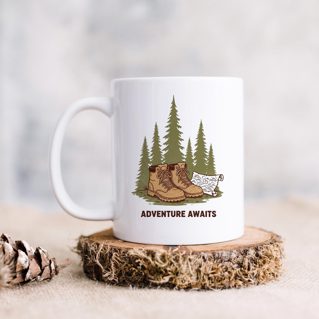White ceramic hiking mug with 'ADVENTURE AWAITS' graphic: boots, pine trees, dog