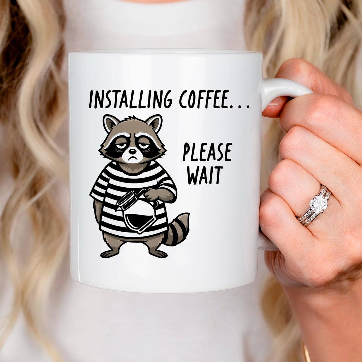 Hand holding white ceramic coffee mug with cartoon raccoon and 'Installing coffee... Please wait'.