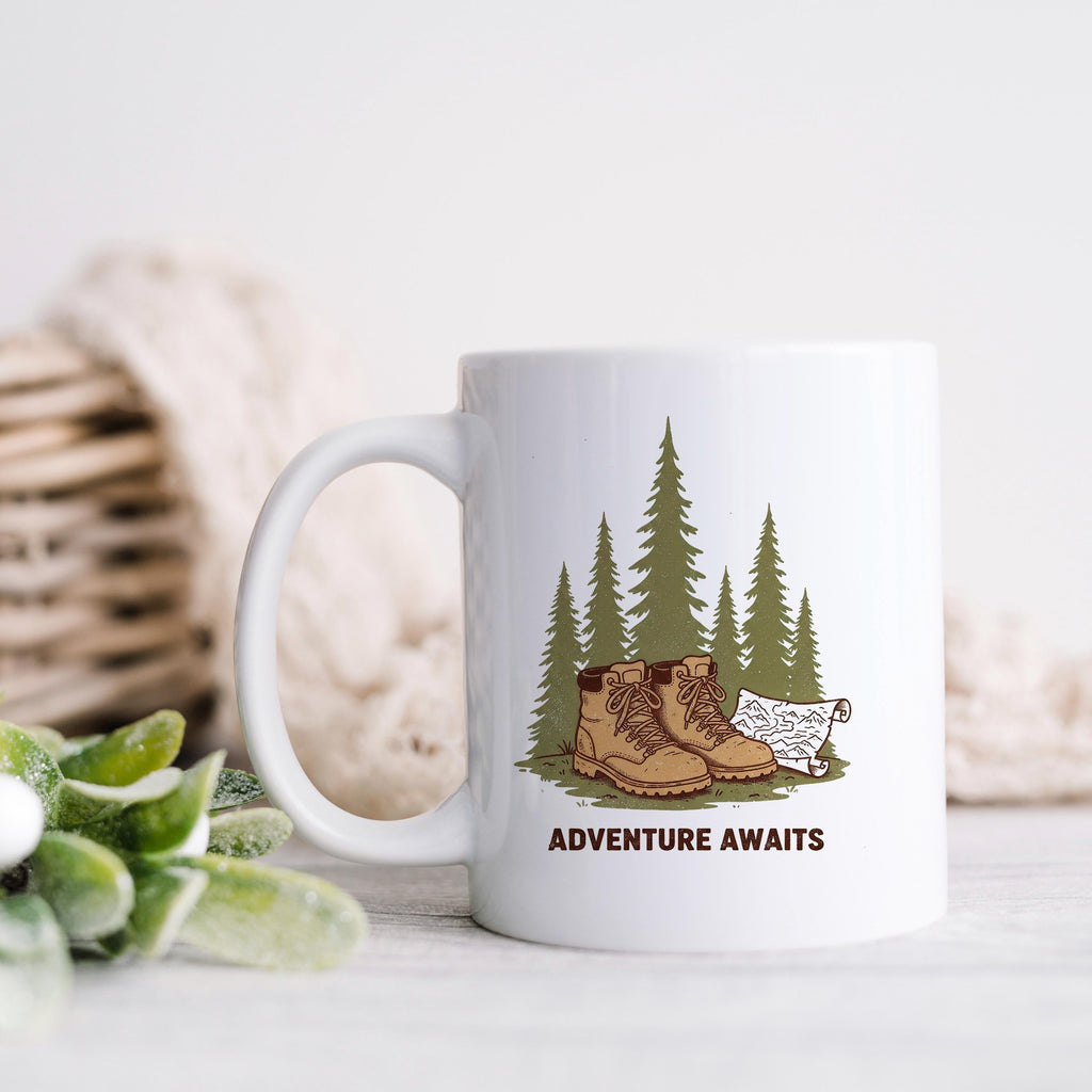 White ceramic mug with 'ADVENTURE AWAITS' graphic of boots, pine trees, and dog