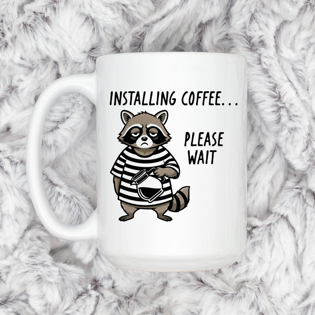 White ceramic coffee mug with cartoon raccoon and 'Installing coffee... Please wait' text.