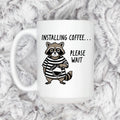 White ceramic coffee mug with cartoon raccoon and 'Installing coffee... Please wait' text.