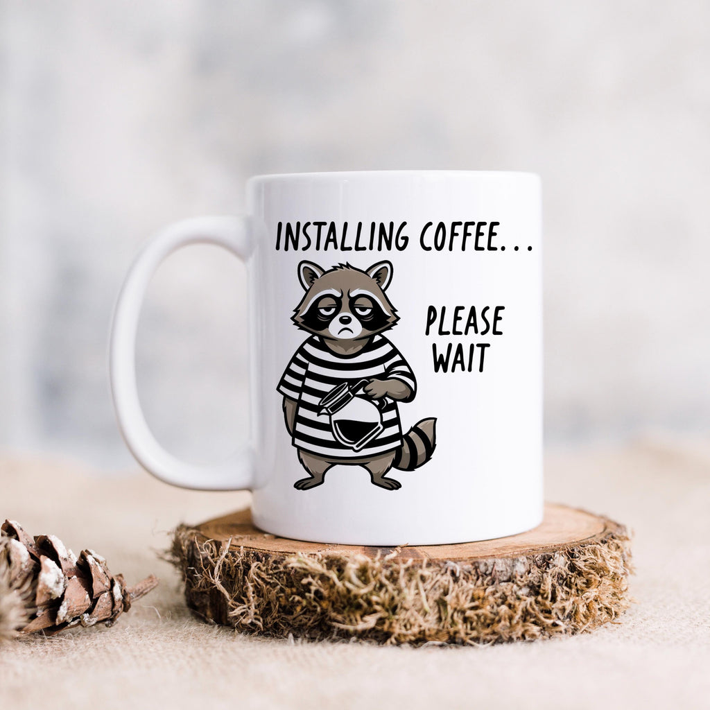 White ceramic coffee mug with cartoon raccoon and 'Installing coffee... Please wait' graphic.
