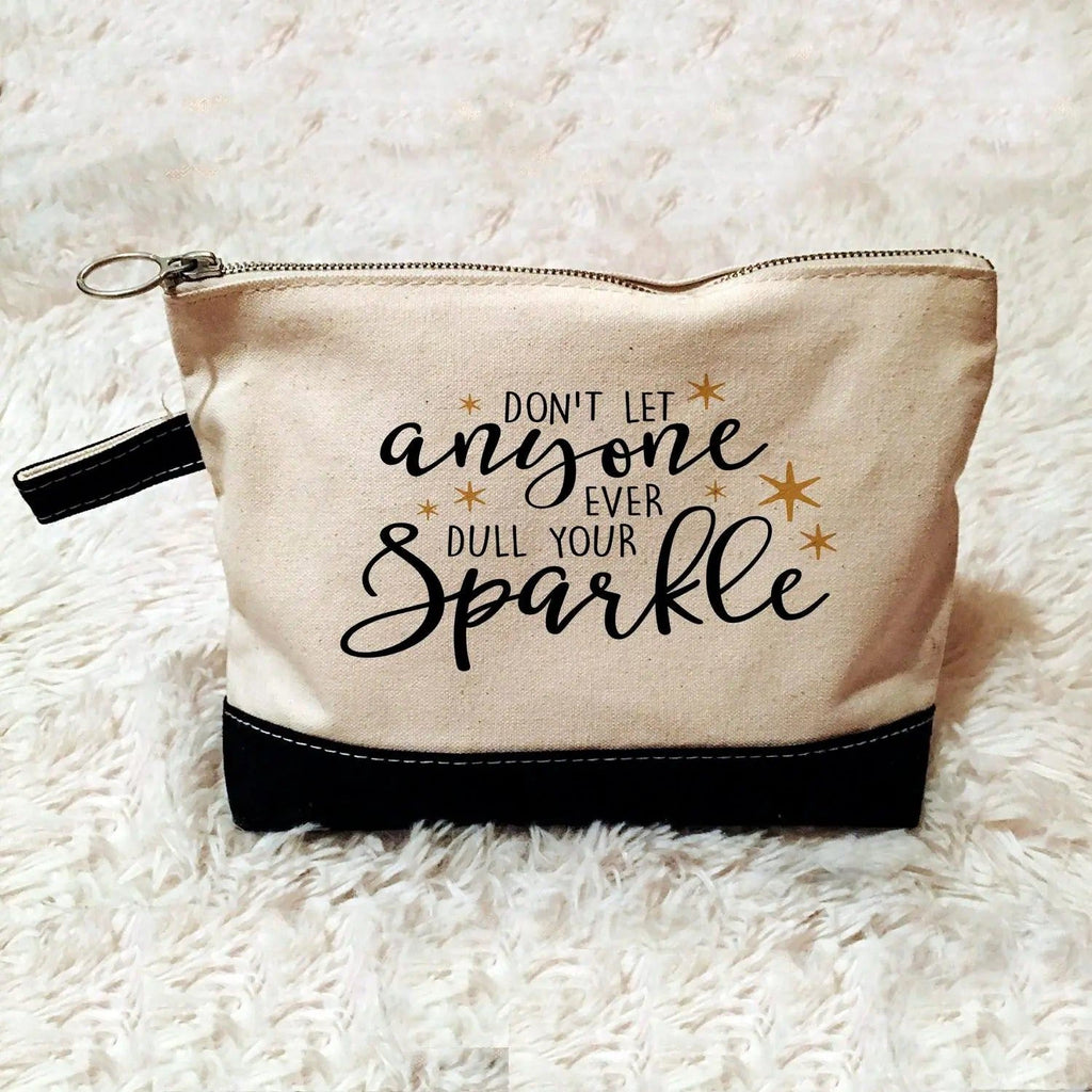 Beige canvas zippered pouch with black bottom and gold slogan 'anyone ... Sparkle'.
