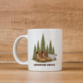White ceramic hiking mug with 'ADVENTURE AWAITS' graphic: boots, pines, and dog