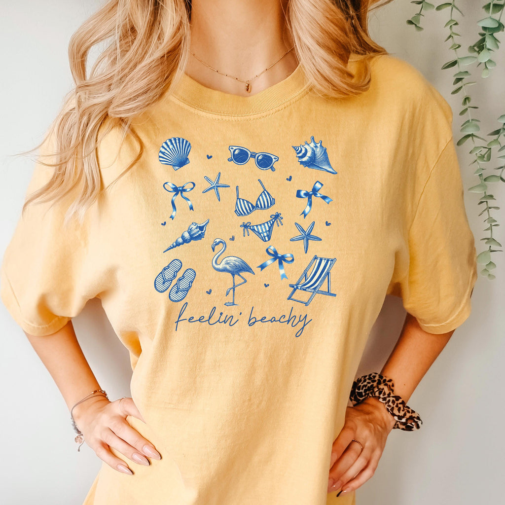 Mustard beach graphic unisex tee with blue shells, flamingos, and “feelin’ beachy” print