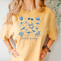 Mustard beach graphic unisex tee with blue shells, flamingos, and “feelin’ beachy” print