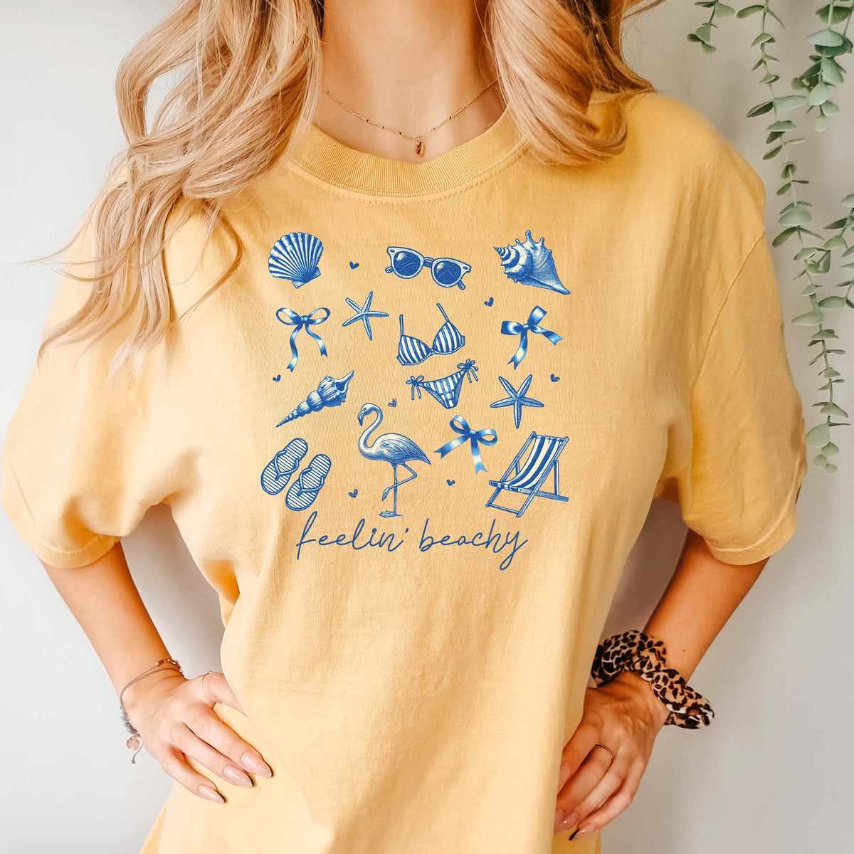 Mustard beach graphic unisex tee with blue shells, flamingos, and “feelin’ beachy” print