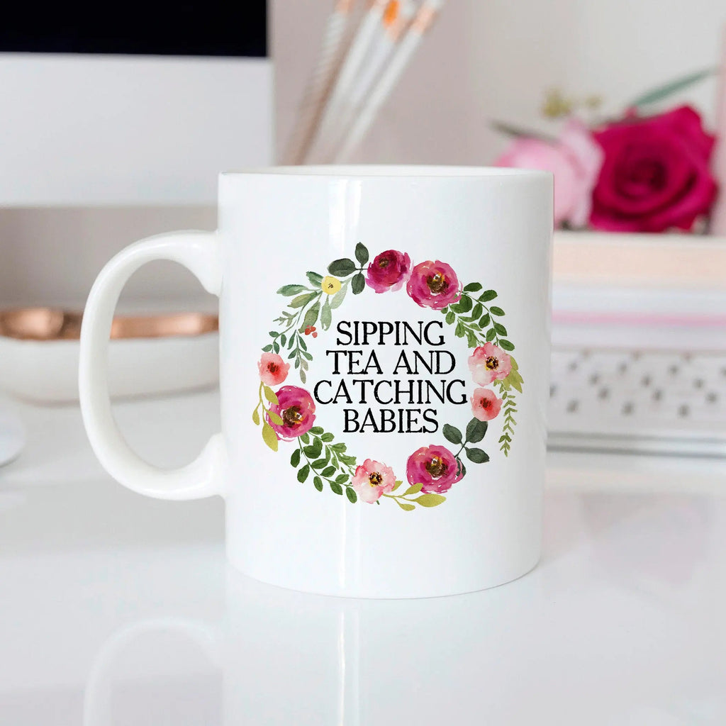 White ceramic mug with floral wreath and text: “SIPPING TEA AND CATCHING BABIES”.