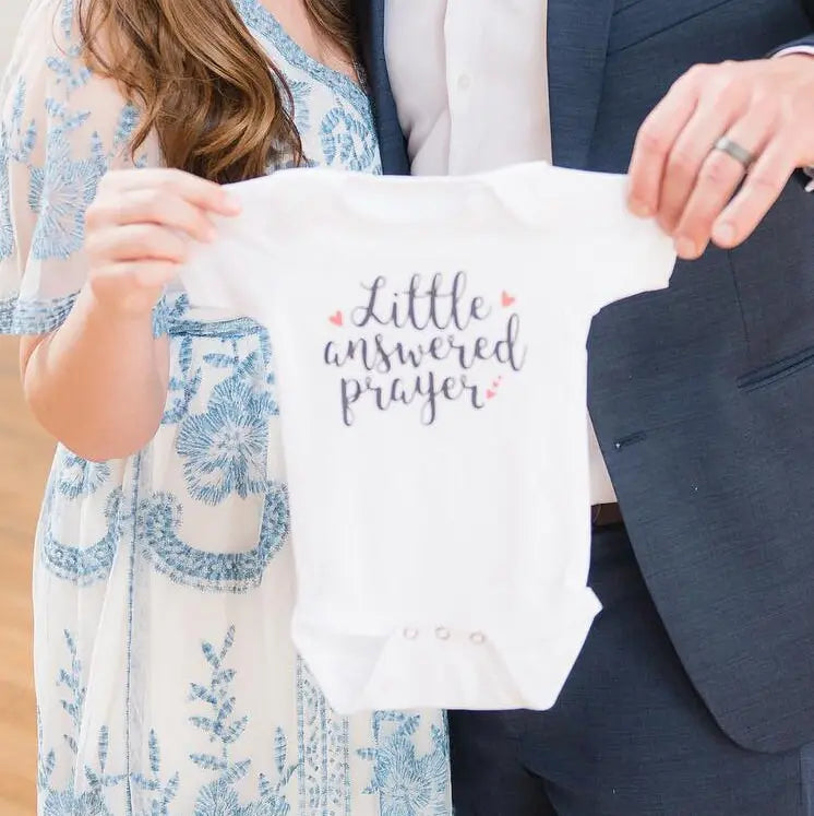 White long-sleeve baby bodysuit with “Little answered prayer” lettering, held up.