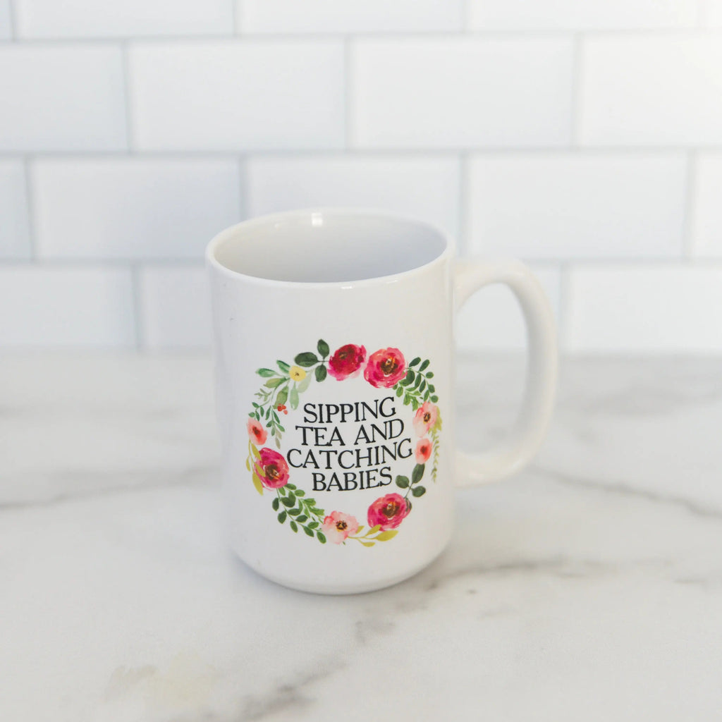 White ceramic mug with floral wreath and “SIPPING TEA AND CATCHING BABIES”.