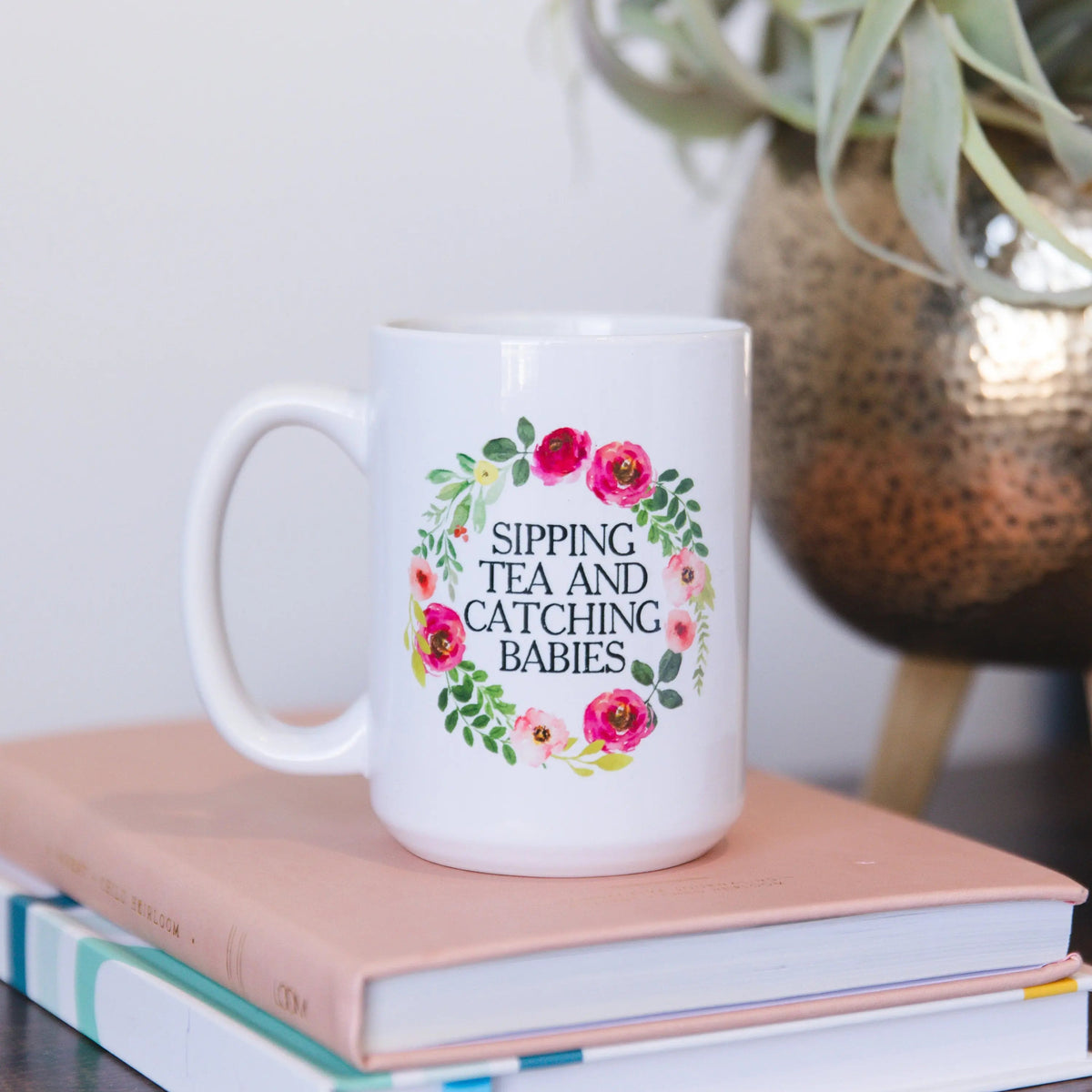 White ceramic mug with floral wreath text: “SIPPING TEA AND CATCHING BABIES”.