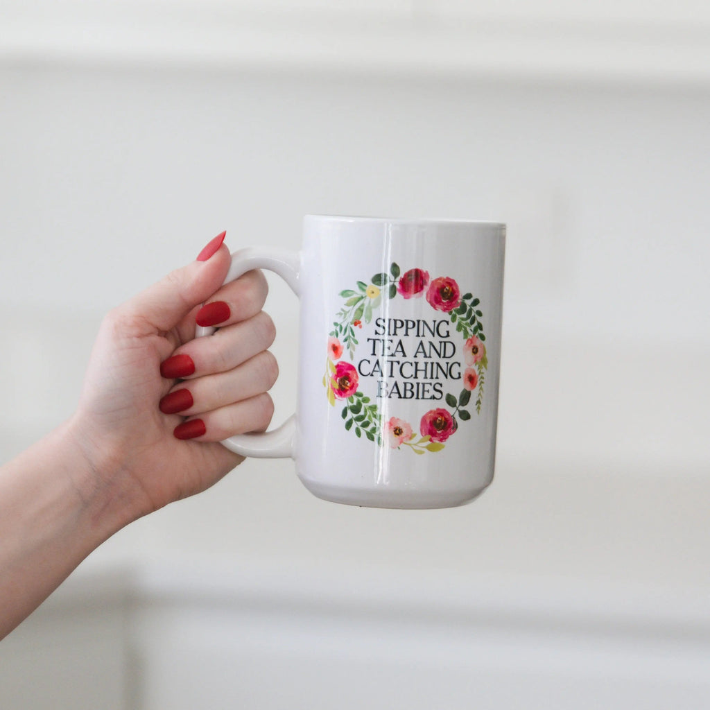 White ceramic mug with floral wreath text: “SIPPING TEA AND CATCHING BABIES”.