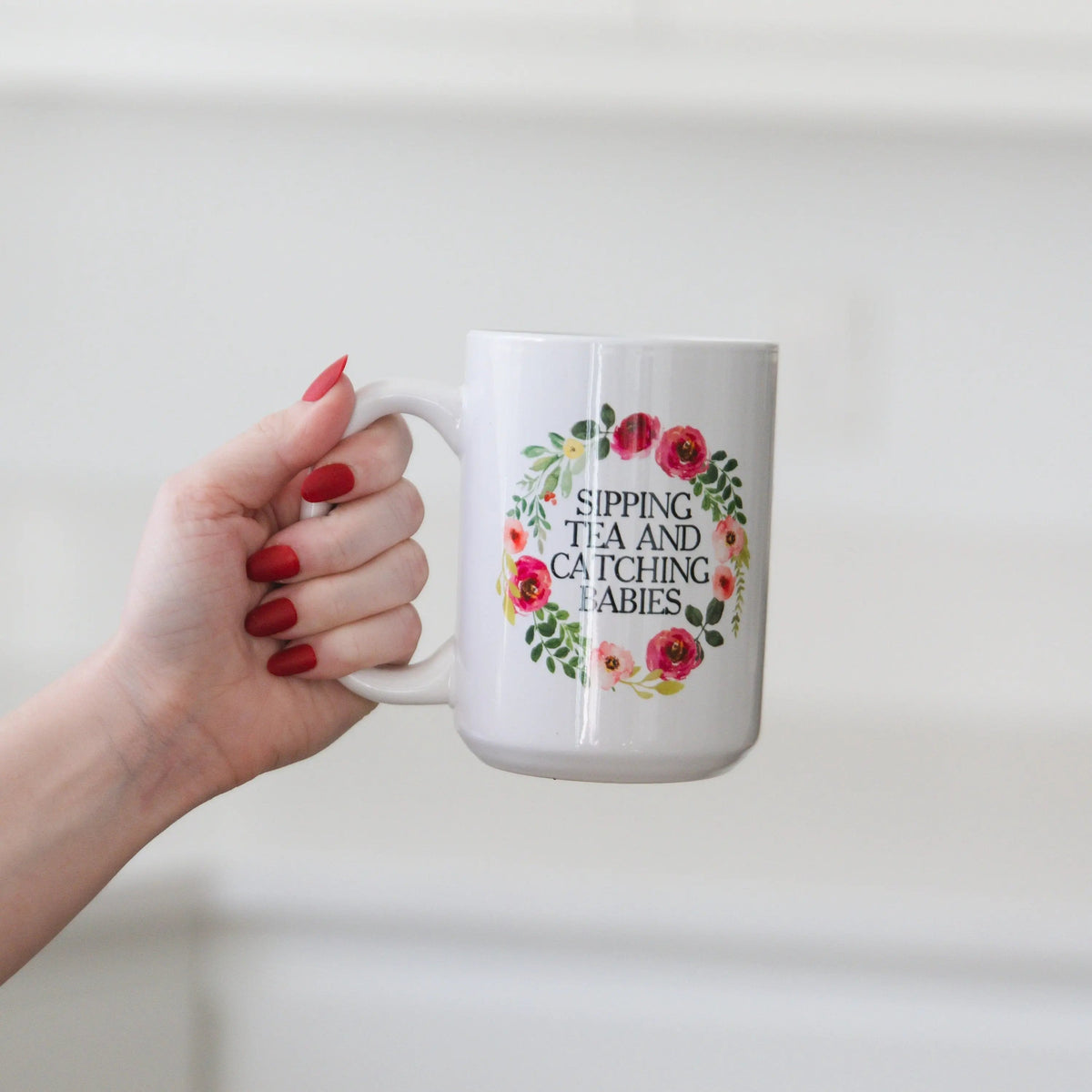 White ceramic mug with floral wreath text: “SIPPING TEA AND CATCHING BABIES”.