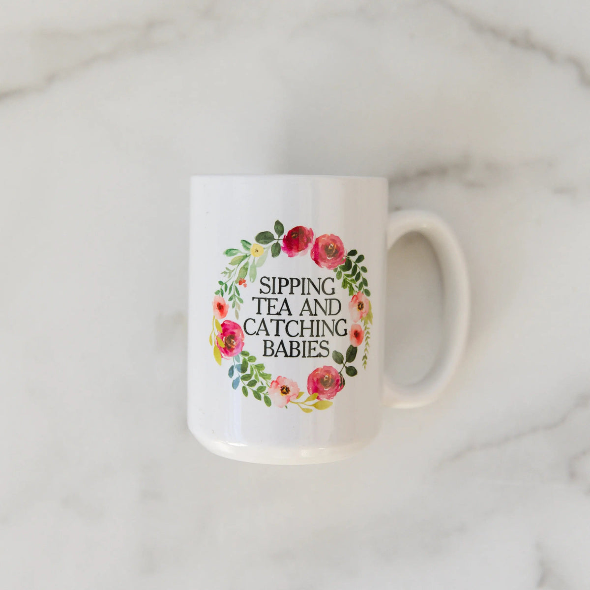 White ceramic mug with floral wreath text: “SIPPING TEA AND CATCHING BABIES”.