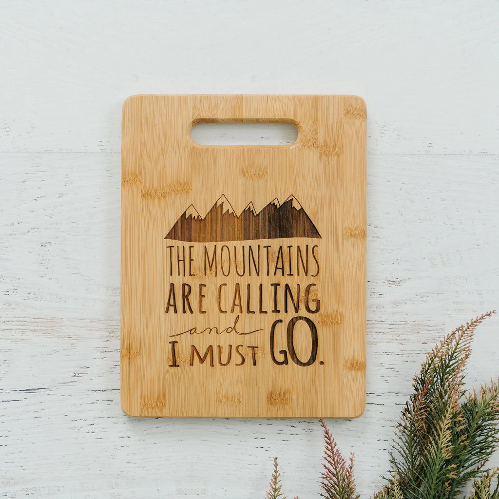 Bamboo cutting board with handle cutout and mountain quote text "The mountains are calling"
