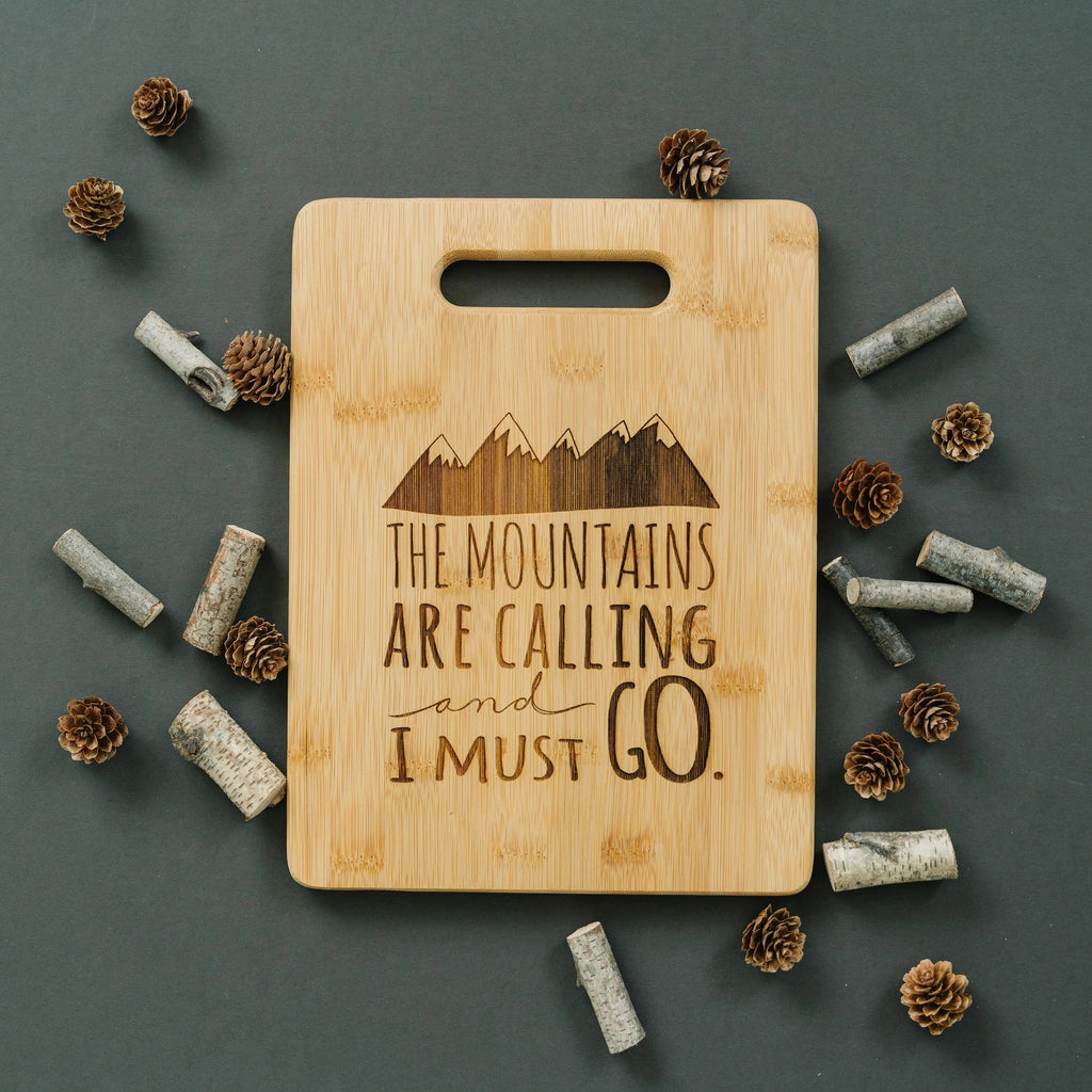 Bamboo cutting board with handle, mountain skyline design, and quote "I must go"