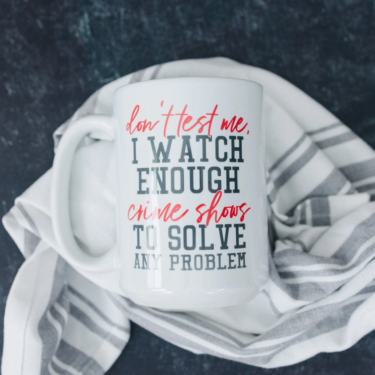 White coffee mug with 'I WATCH ENOUGH...' crime shows quote on a striped blanket