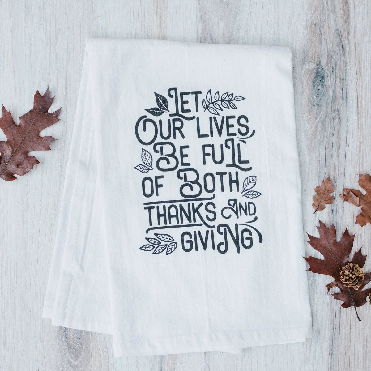 White Thanksgiving kitchen towel laid on wood with black quote and leaf accents