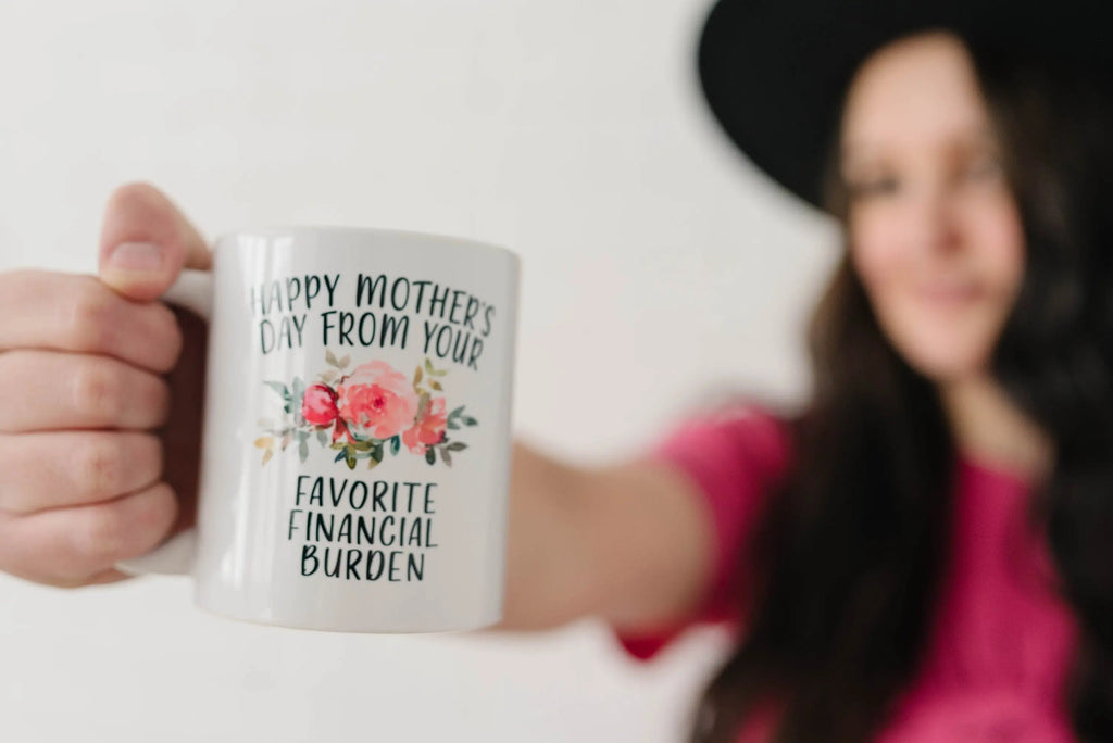 Hand holding white ceramic coffee mug with floral “Happy Mother’s Day” and “Favorite Financial Burden”
