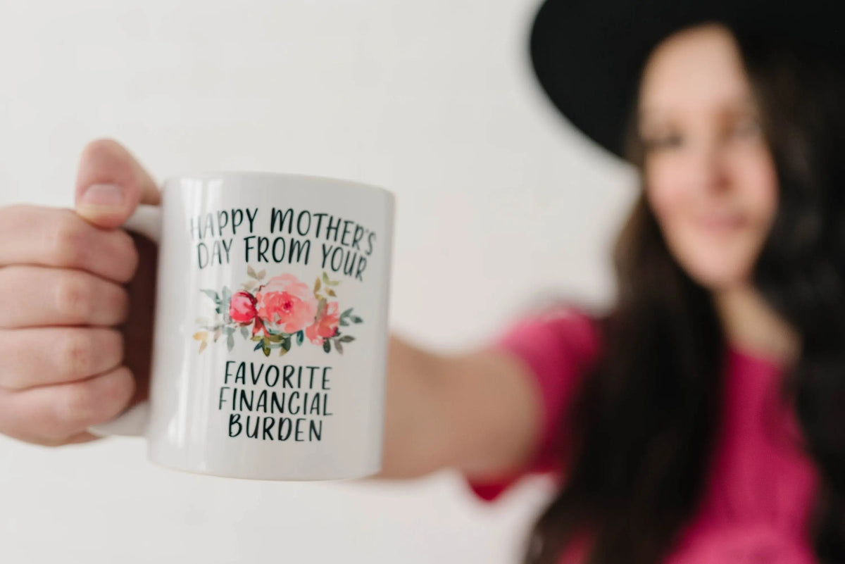 Hand holding white ceramic coffee mug with floral “Happy Mother’s Day” and “Favorite Financial Burden”