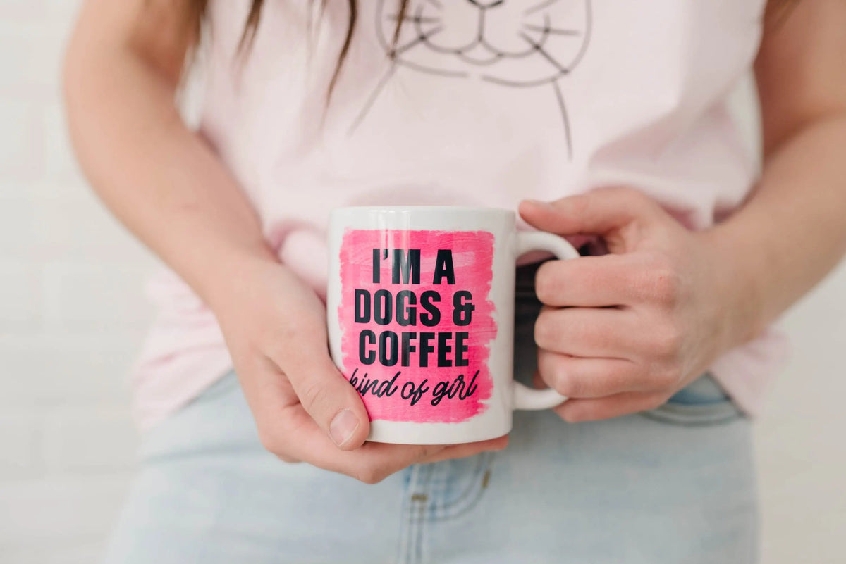 White coffee mug in hands with pink logo: “I'm a DOGS & COFFEE kind of girl”