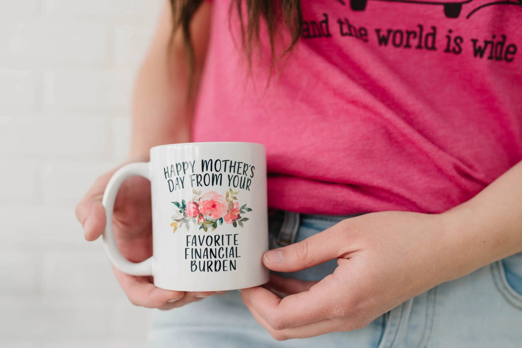 Hand holding white ceramic coffee mug with “Happy Mother’s Day” and “Favorite Financial Burden”